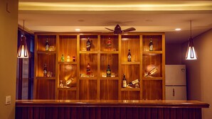 A bar area with wooden shelves holding various bottles and glasses, a ceiling fan, and a refrigerator. - Safari Adventure Lodge (Sauraha)