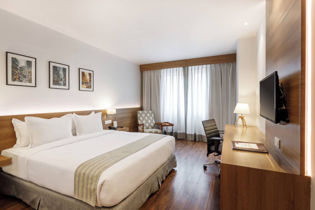 A hotel room with a large bed, a desk with a lamp, a TV, and three framed pictures on the wall.