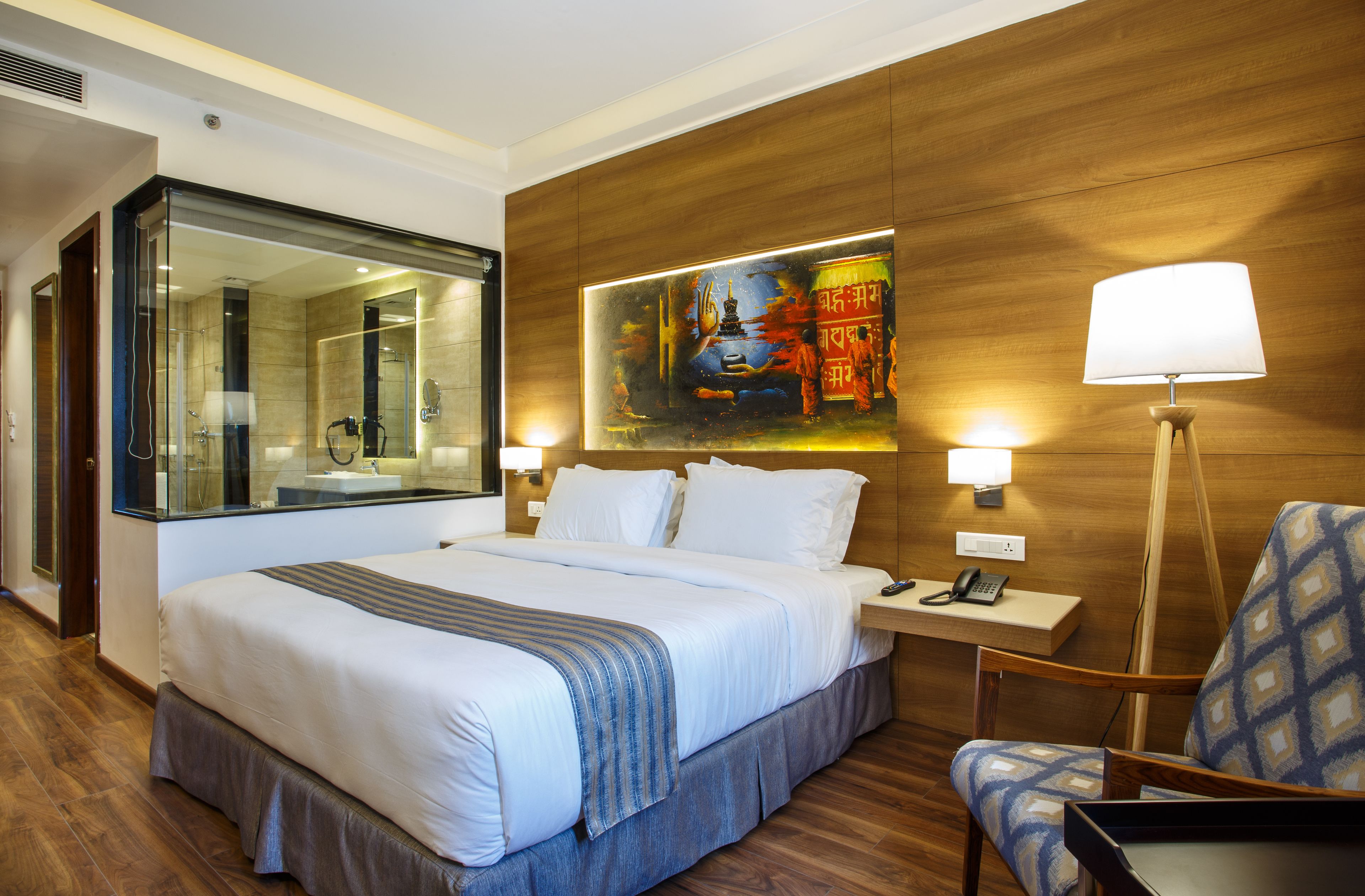A hotel room with a large bed, a bedside table with a lamp, a chair, a painting on the wall, and a mirror.