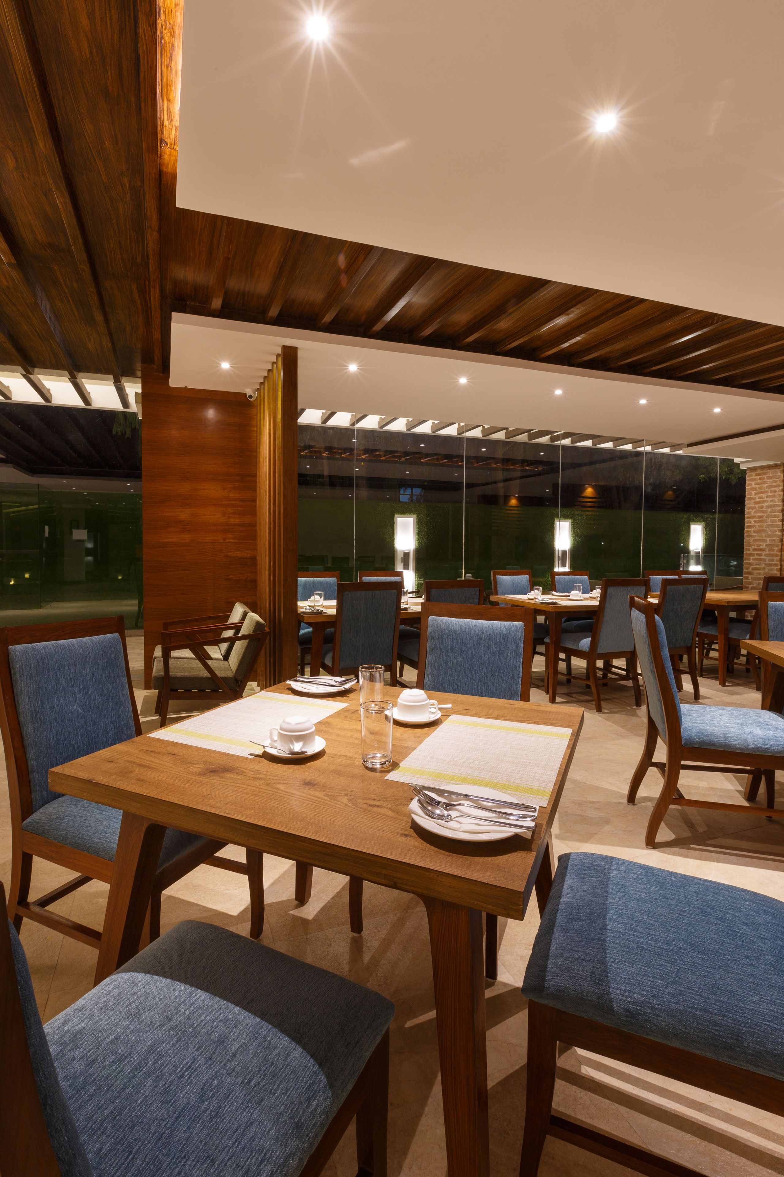 A restaurant with wooden tables and blue upholstered chairs, featuring a modern interior with a glass wall and wooden ceiling beams.
