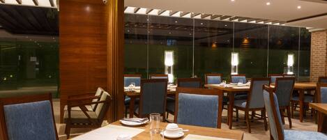 A restaurant with wooden tables and blue upholstered chairs, featuring a modern interior with a glass wall and wooden ceiling beams.
