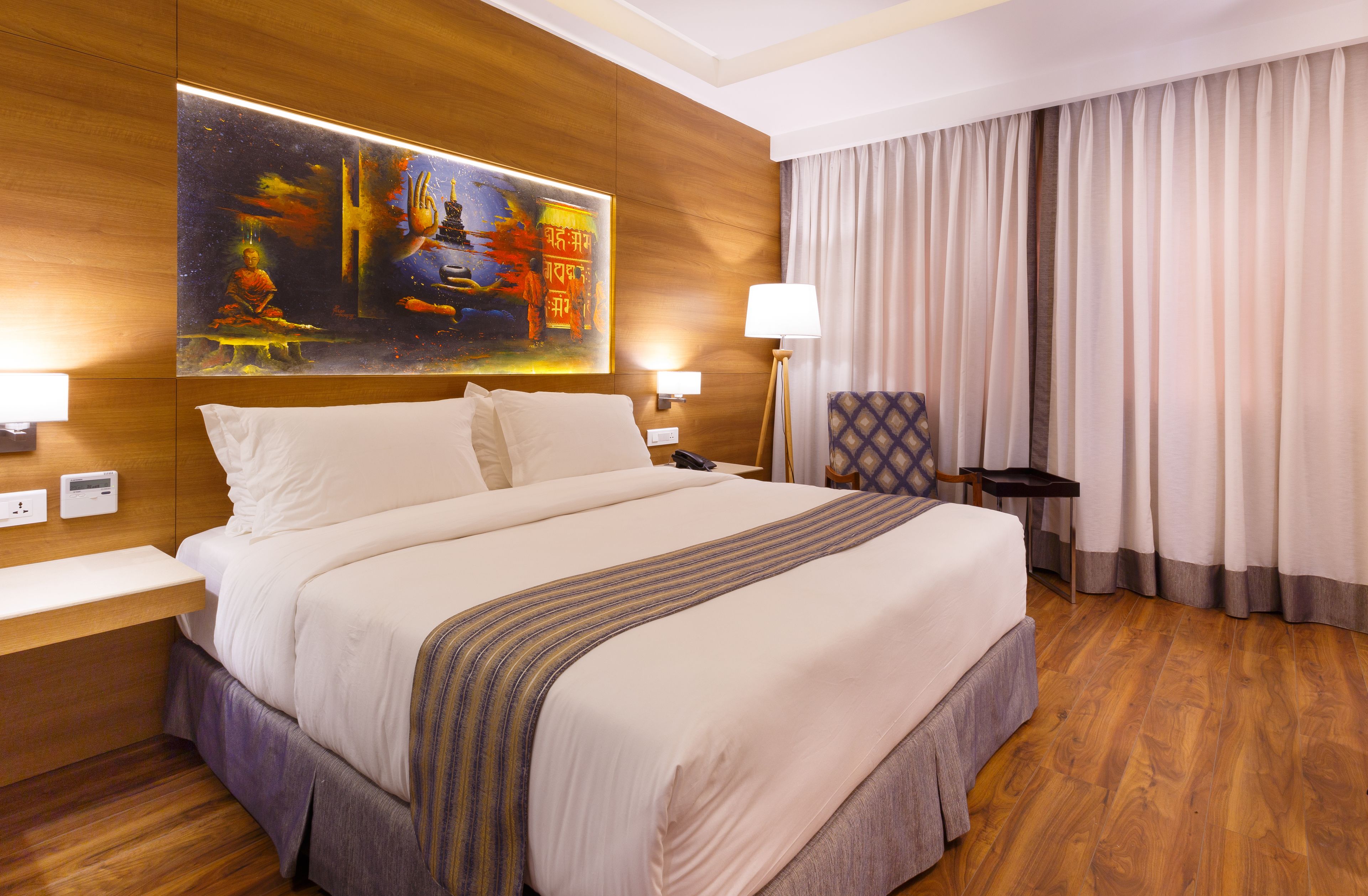 A hotel room with a large bed, wooden flooring, a painting on the wall, a desk, and a chair.