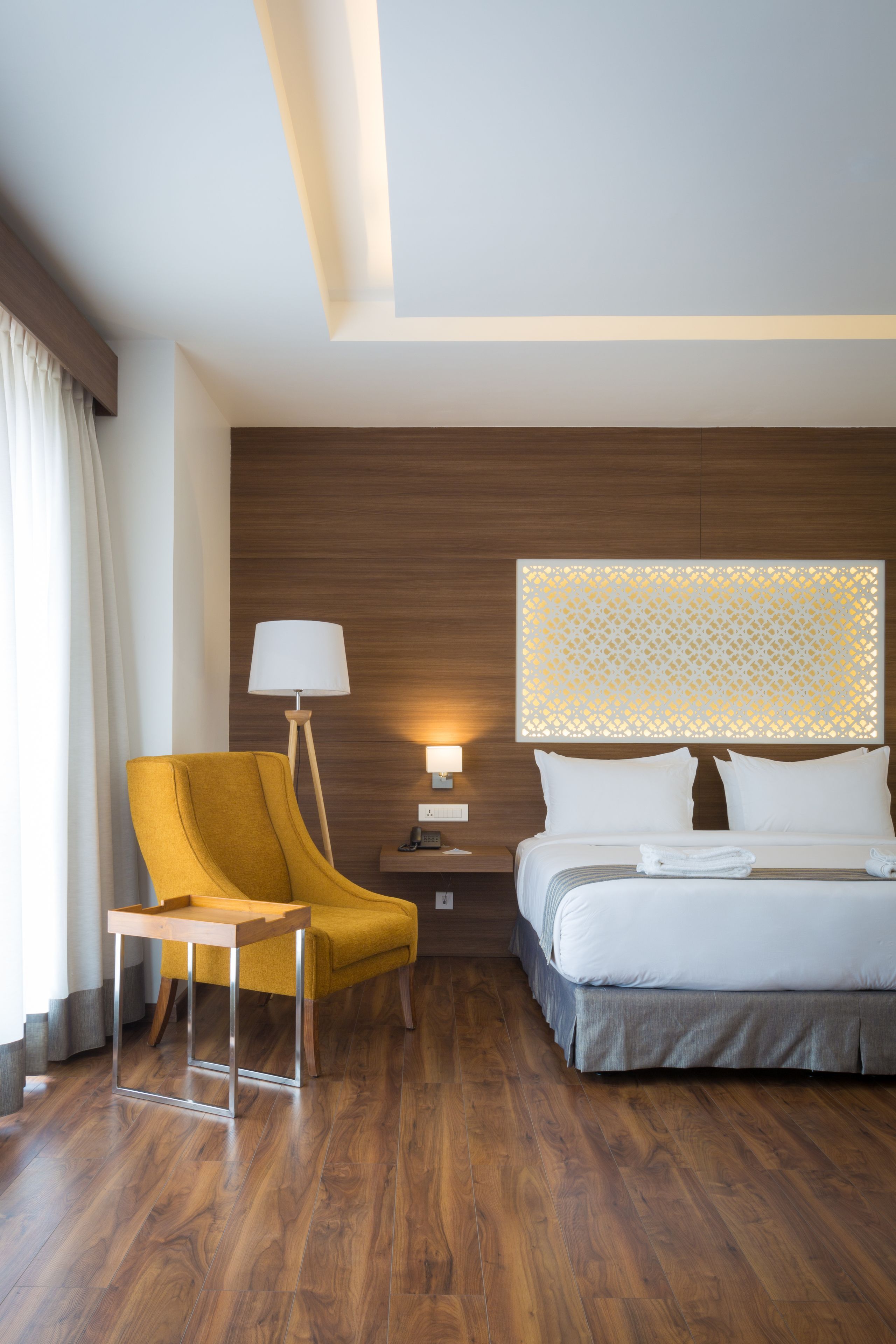Una habitación de hotel con suelo de madera, un sillón amarillo, una cama con sábanas blancas, una mesita de noche con lámpara y un panel decorativo en la pared.