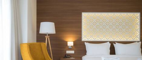 Una habitación de hotel con suelo de madera, un sillón amarillo, una cama con sábanas blancas, una mesita de noche con lámpara y un panel decorativo en la pared.