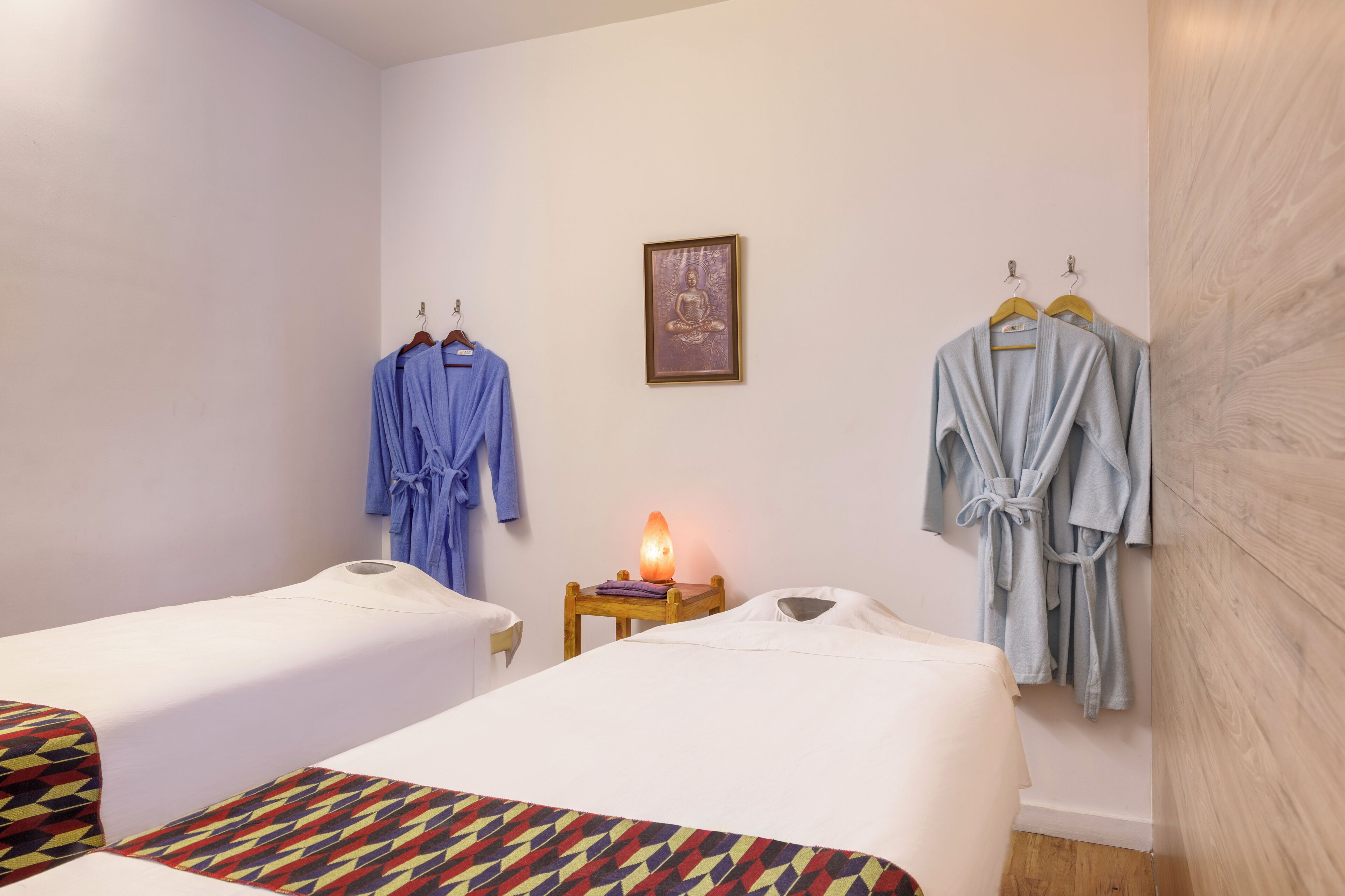 A hotel room with two beds, a small table with a lamp, and two robes hanging on the wall.