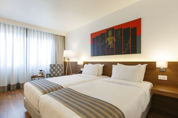 A hotel room with two beds, a large painting on the wall, a lamp, and a window with curtains.