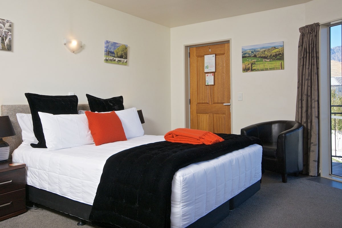 A hotel room with a bed, a desk with a lamp, a chair, and artwork on the walls.