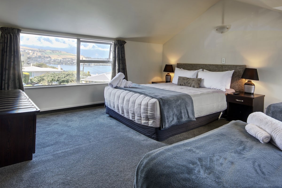 A hotel room with a large bed, two bedside tables, a desk, and a view of a lake and buildings.