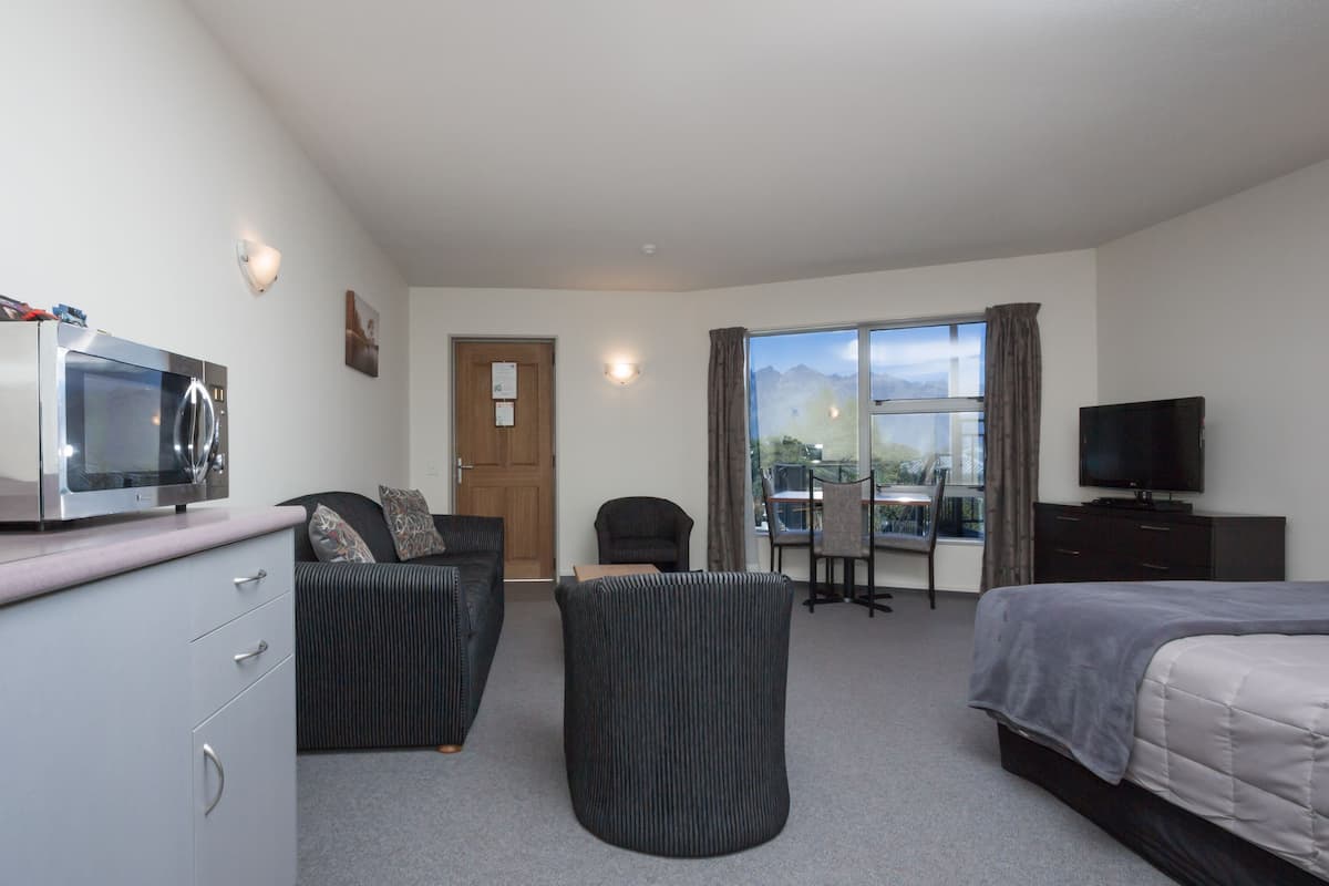 A hotel room with a bed, a desk with a computer, a chair, a sofa, a small table, a television, and a window with a view of mountains.