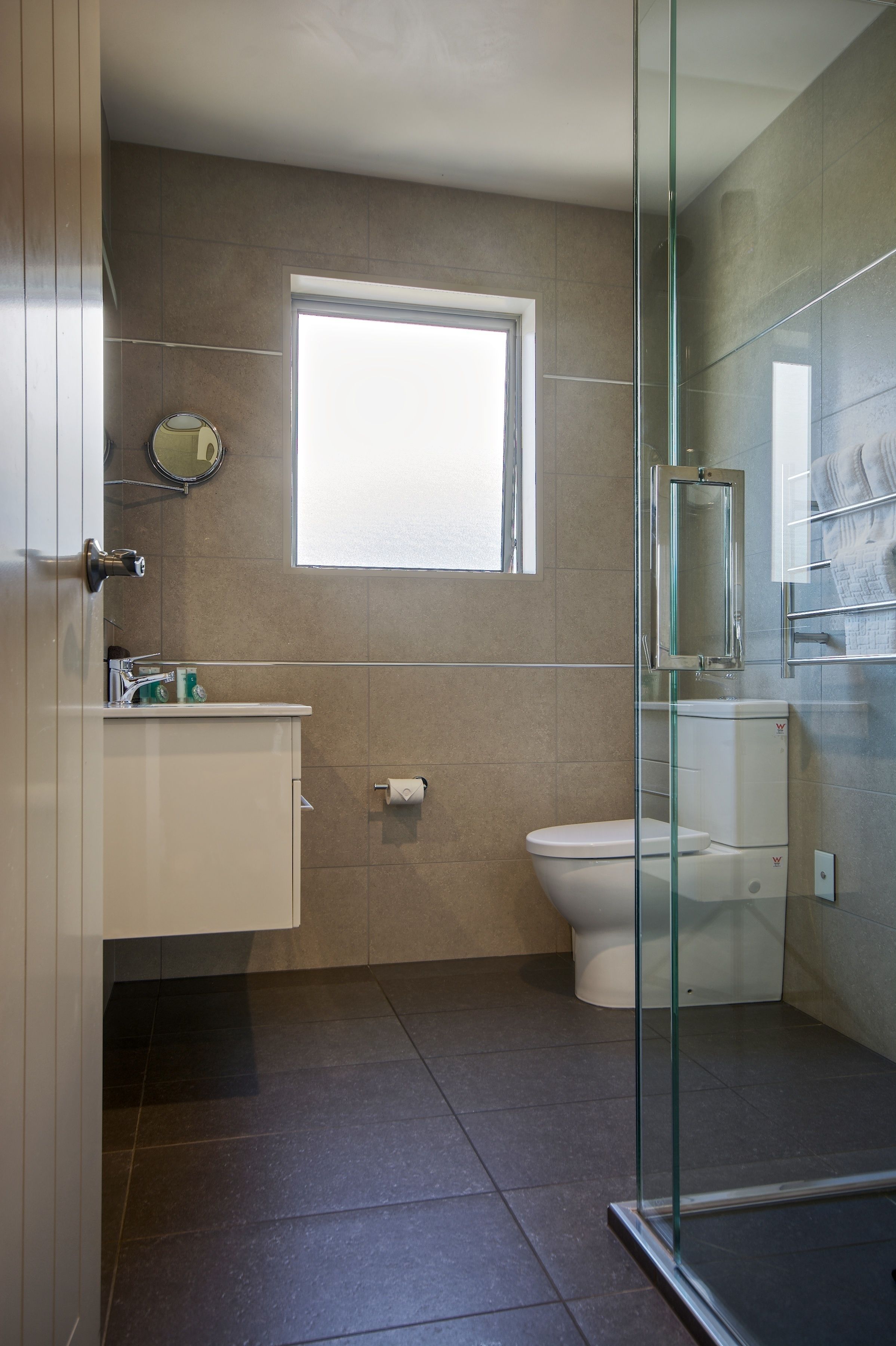 Un baño con inodoro, lavamanos, cabina de ducha de vidrio y ventana que proporciona luz natural.