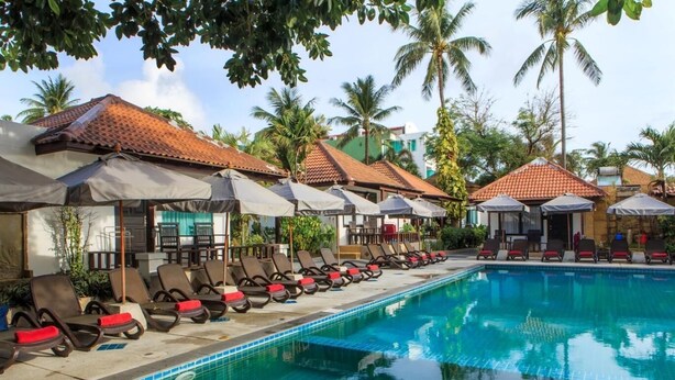 A swimming pool with lounge chairs and umbrellas, surrounded by palm trees and buildings with red-tiled roofs.