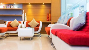 A modern hotel lobby with white and orange sofas, a white coffee table, and wooden shelves with magazines.