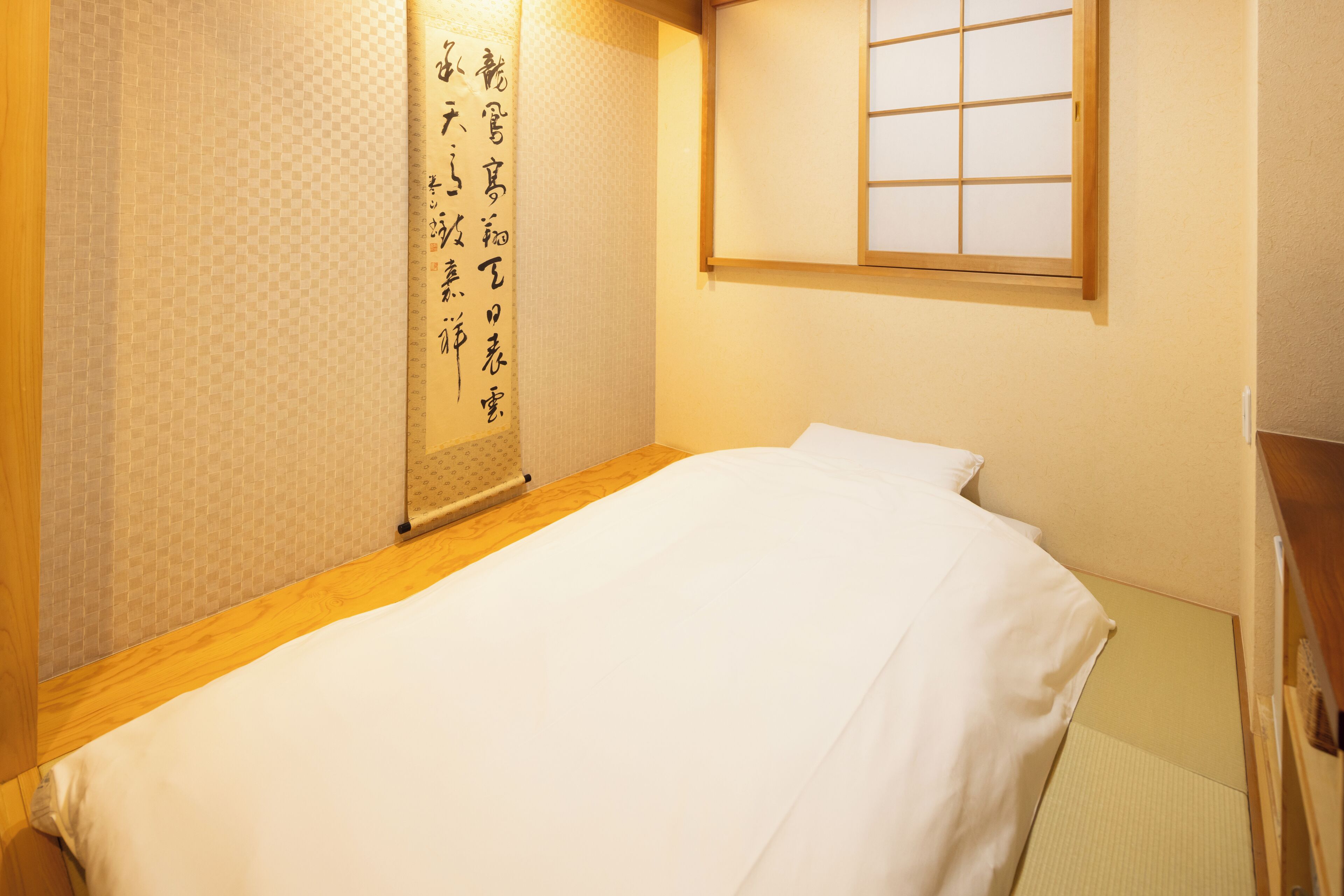 Un lit bien fait dans une pièce dotée d’une tête de lit en bois, d’un paravent japonais orné de calligraphies et d’une fenêtre à carreaux.