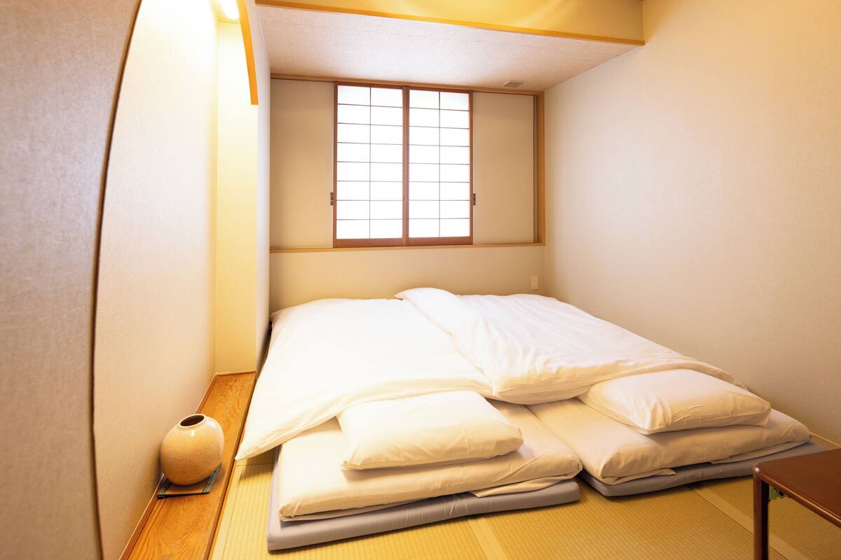 A small, neatly made bed in a room with tatami flooring, a sliding shoji door, and a small table with a vase.