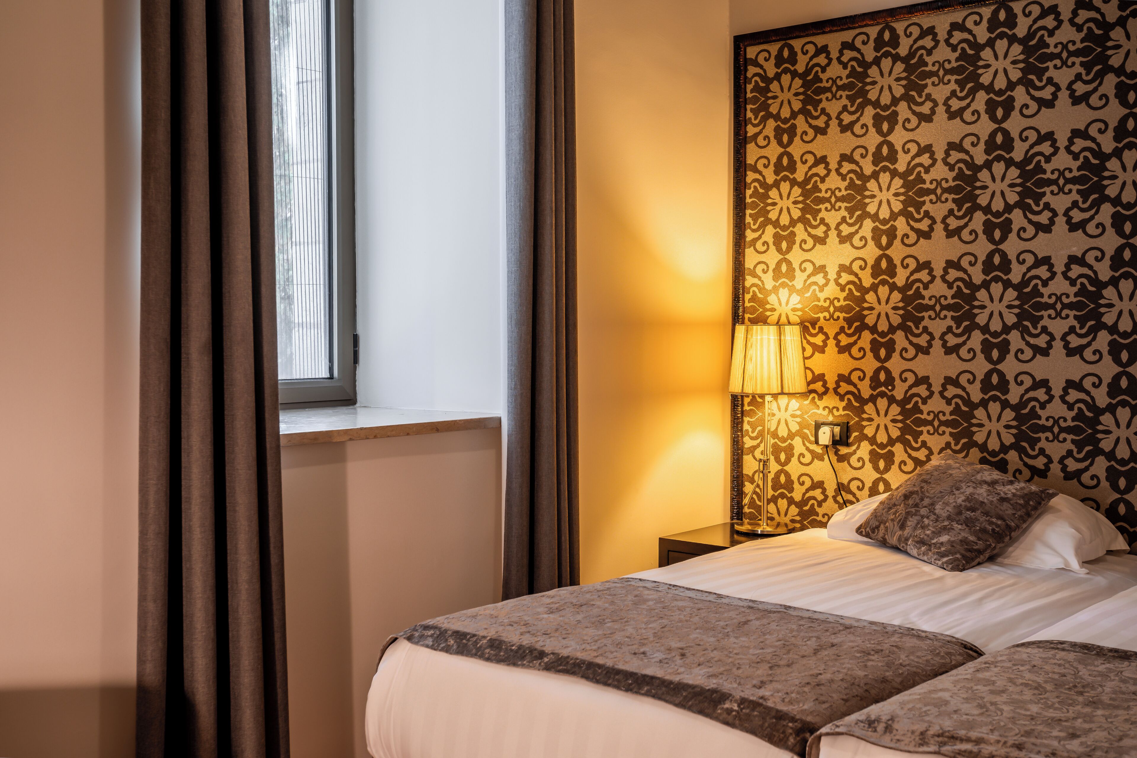 A hotel room with a bed, curtains, a lamp, and a patterned wall.