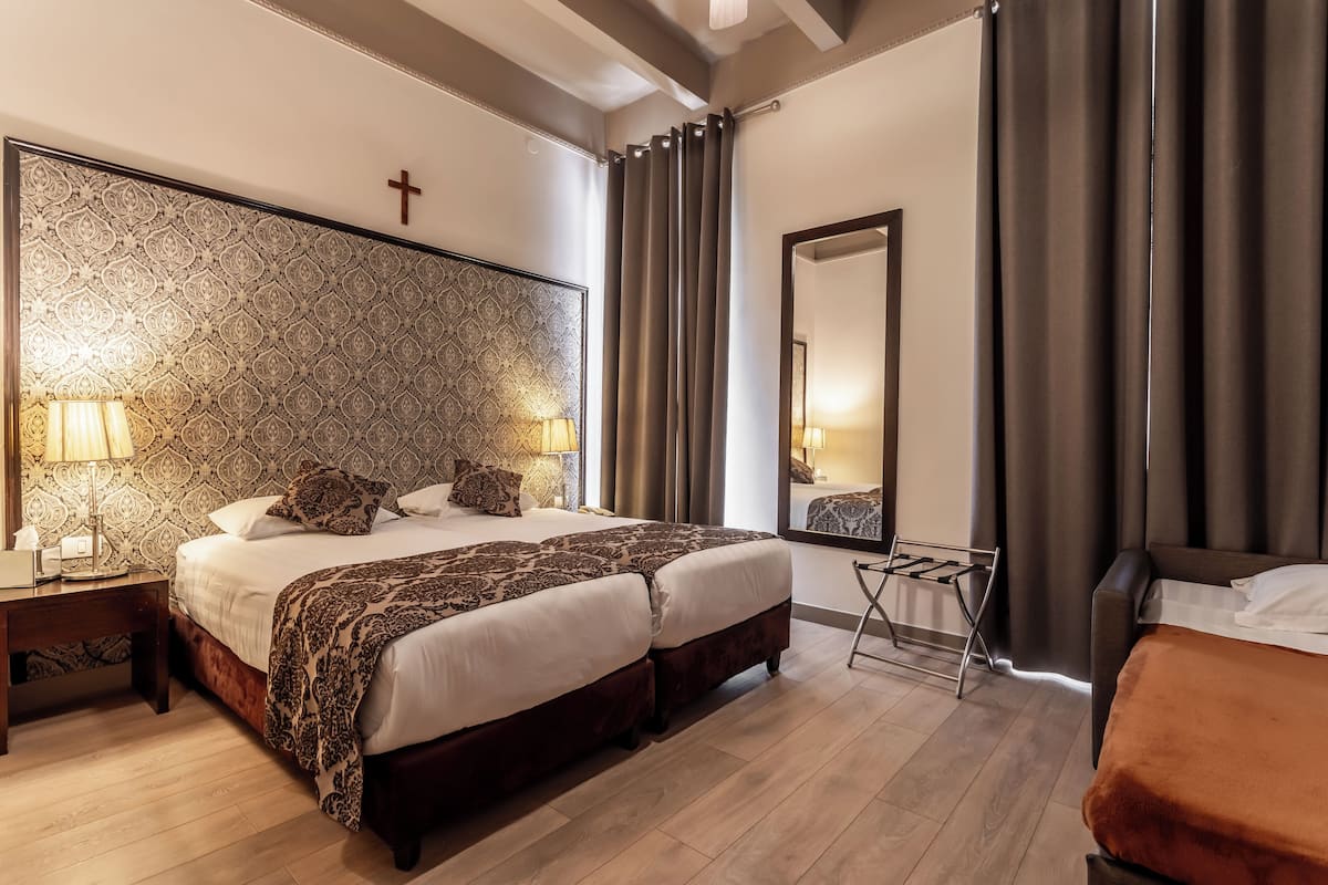 A hotel room with two beds, a large mirror, a cross on the wall, and a patterned headboard.