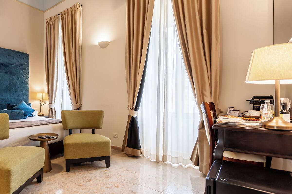 A hotel room with a bed, two chairs, a desk, a lamp, and a cross on the wall.