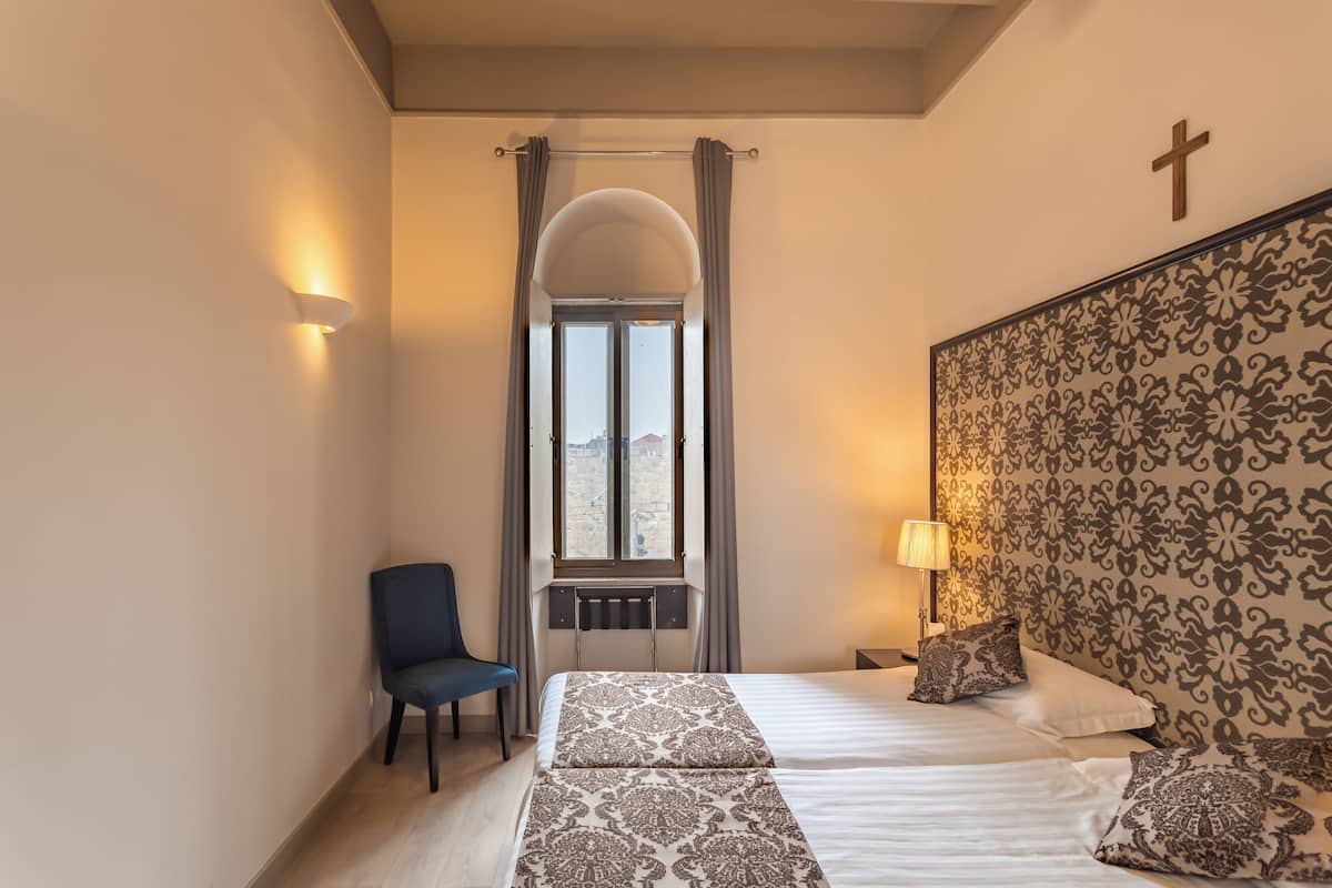 A hotel room with two beds, a chair, a window with curtains, a cross on the wall, and a patterned headboard.
