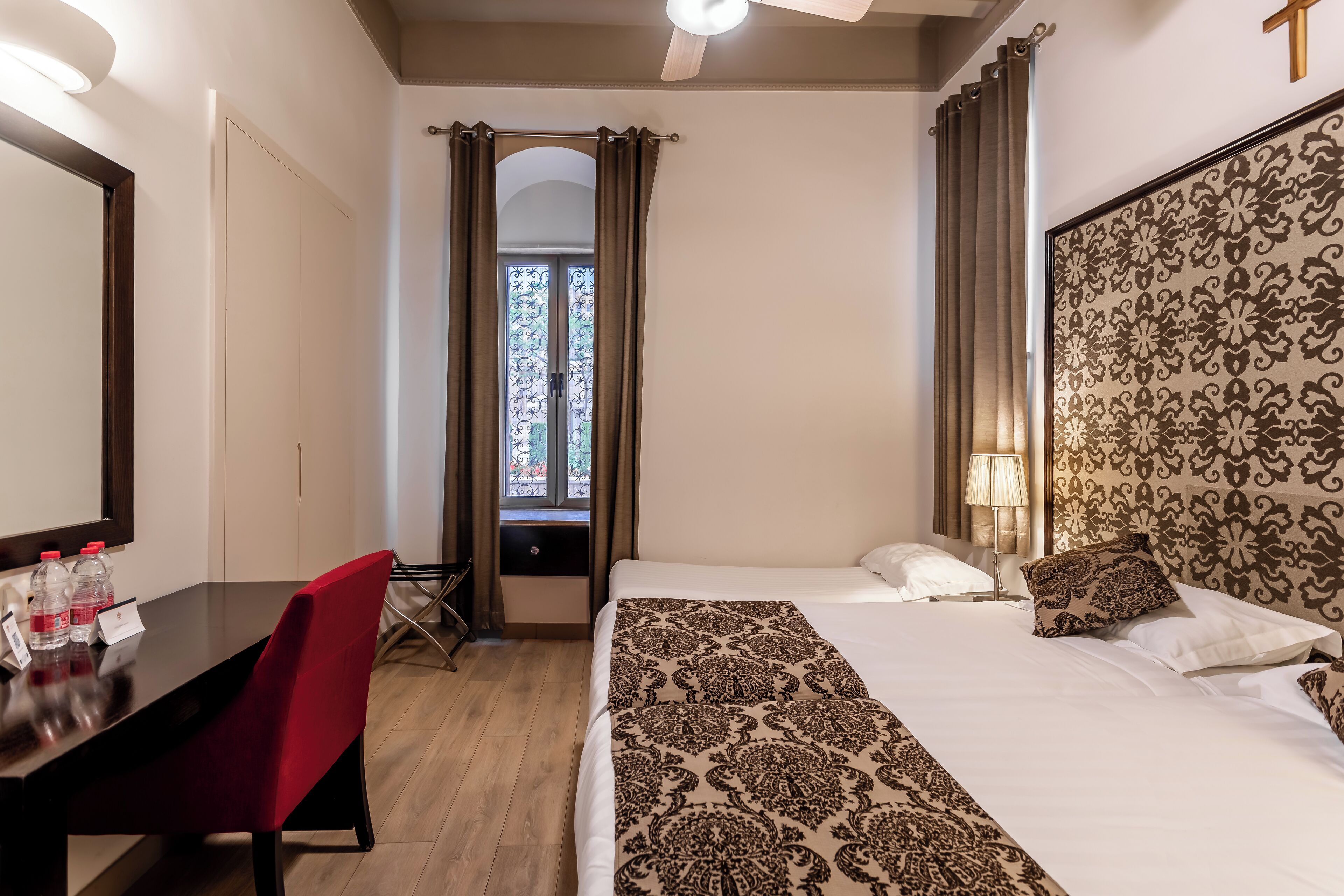 Quarto de hotel com uma cama, uma escrivaninha com cadeira, uma janela com cortinas e um ventilador de teto.