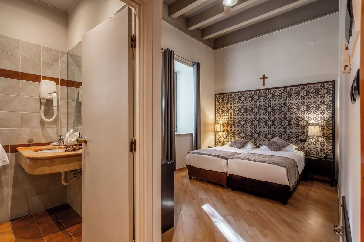 A hotel room with a wooden floor, a bed with a cross on the headboard, a bathroom with a sink and mirror, and a patterned wall.