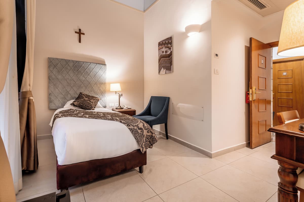 A hotel room with a bed, a chair, a lamp, a cross on the wall, and a door.