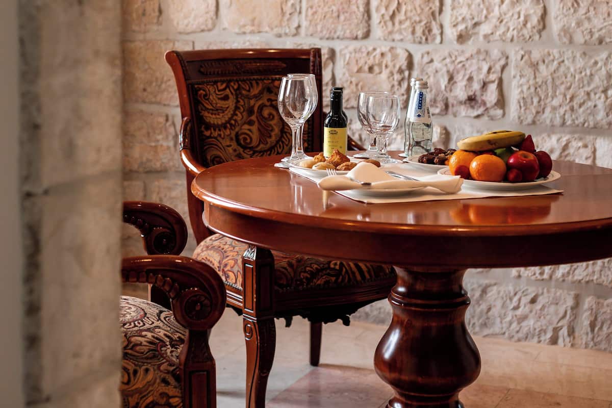 A round wooden table with a plate of fruit and two wine glasses, set against a stone wall.