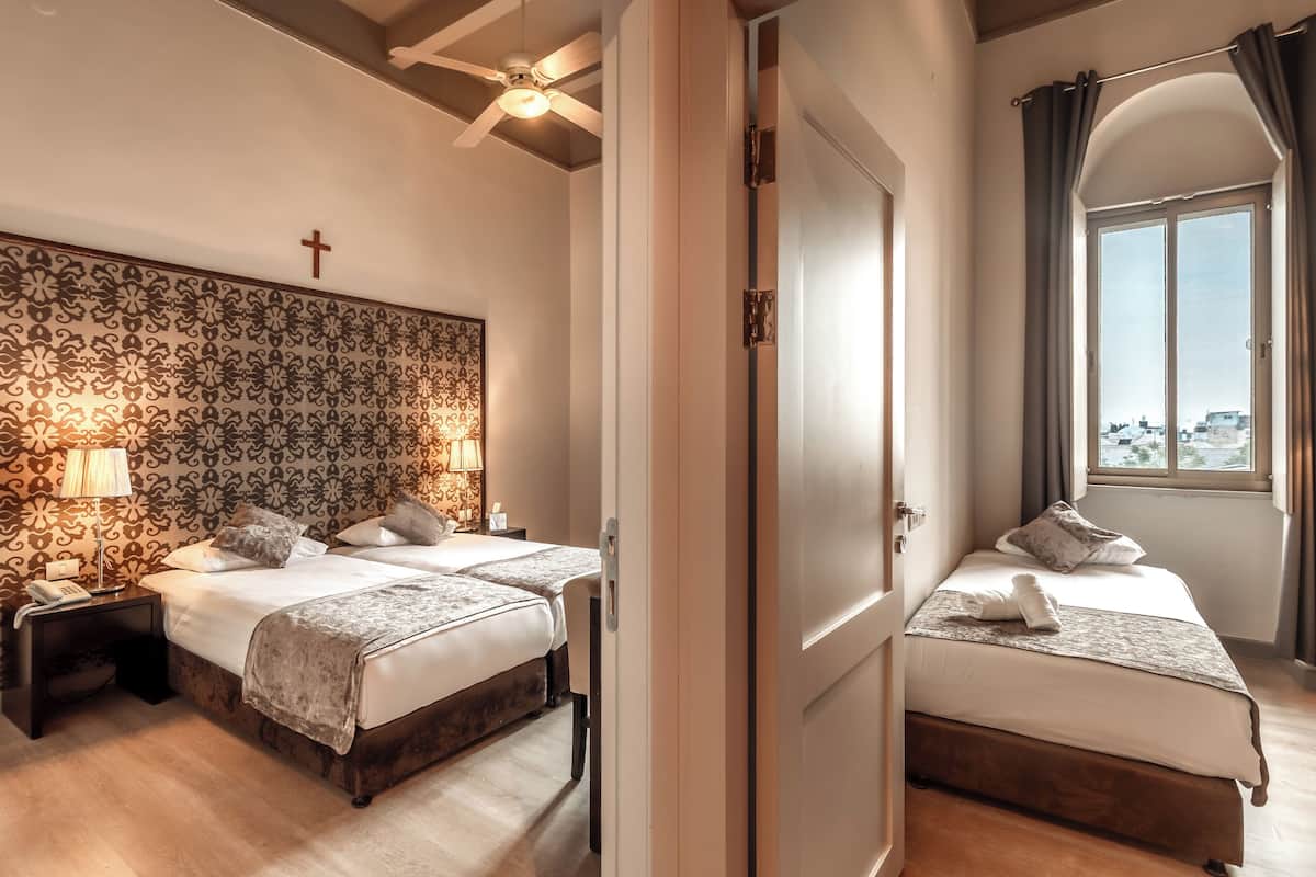 A hotel room with two beds, a patterned headboard, a cross on the wall, a ceiling fan, and a window with curtains.