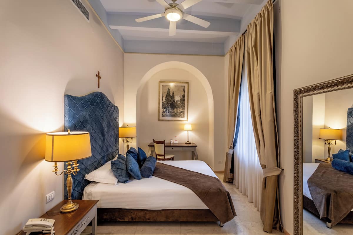 A hotel room with a bed, two bedside lamps, a desk, a chair, a mirror, and a cross on the wall.