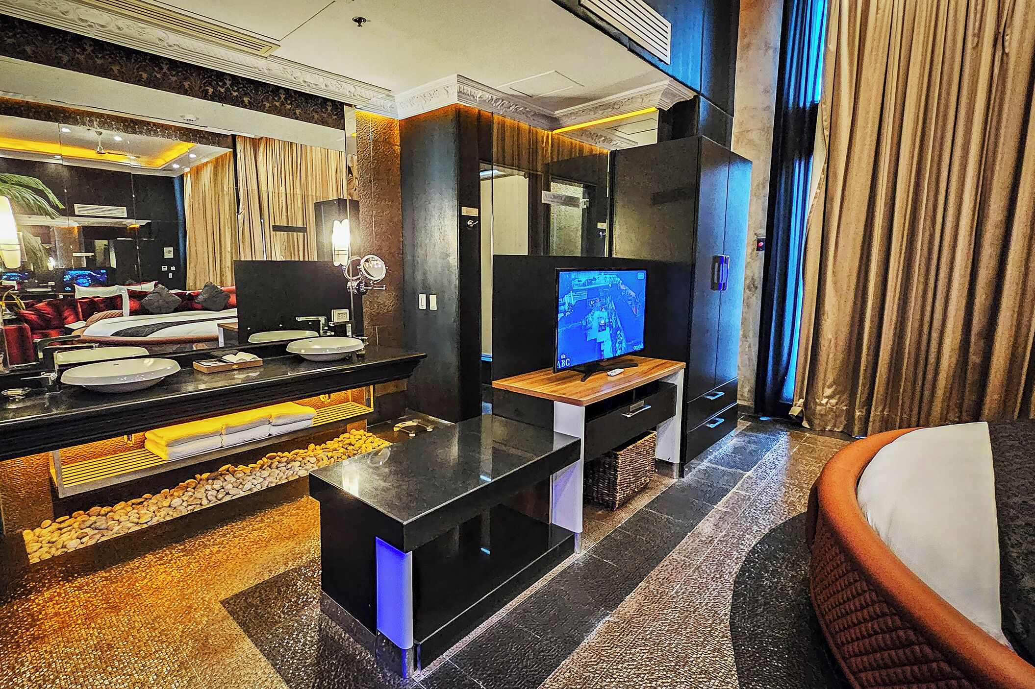 A modern hotel room with a bed, a TV, a desk with a computer, and a large mirror.
