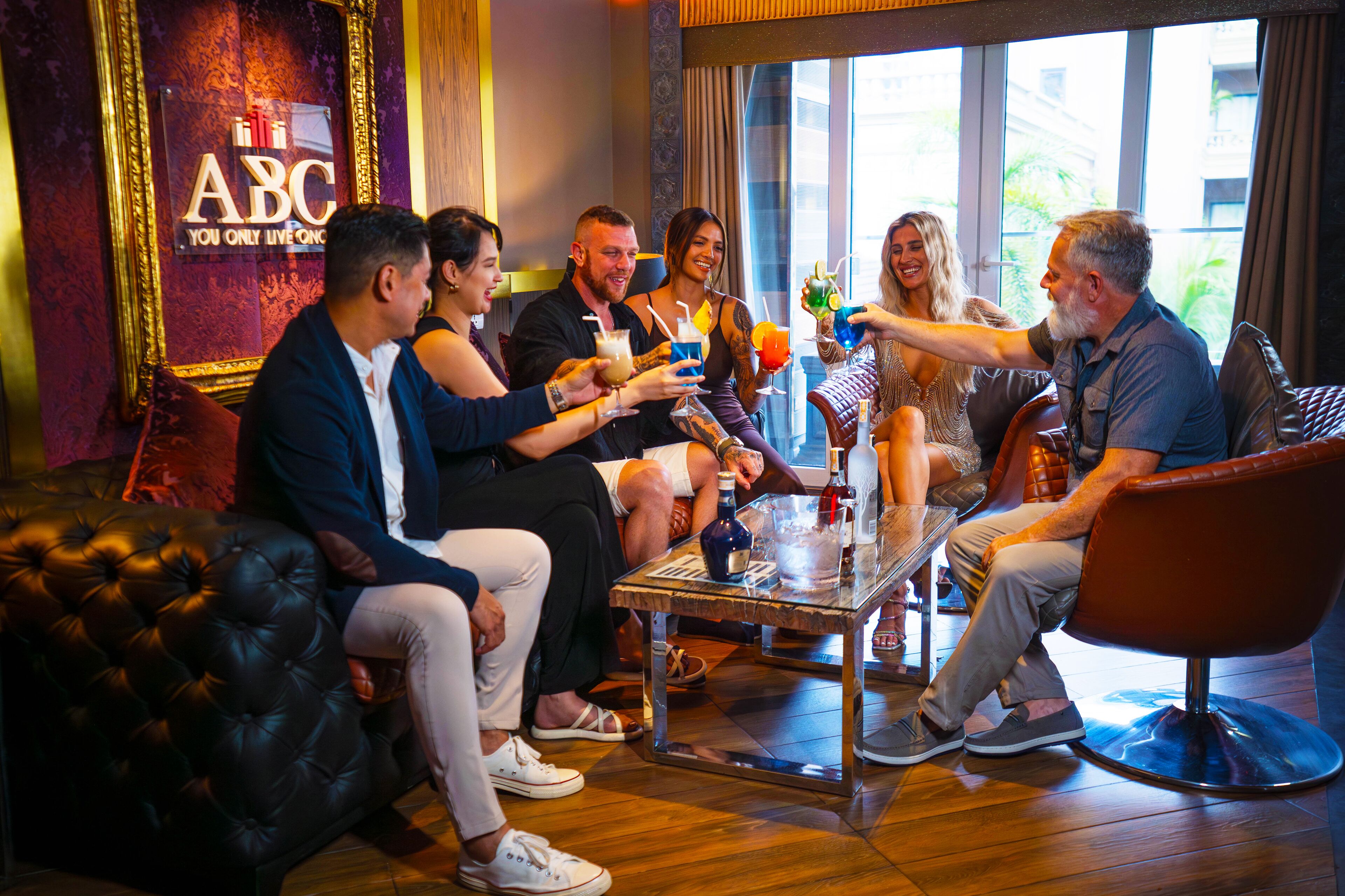 Un grupo de personas sentadas alrededor de una mesa en una habitación con un ventanal grande, un sofá de cuero y un cartel que dice "ABC YOU ONLY LIVE ONCE".