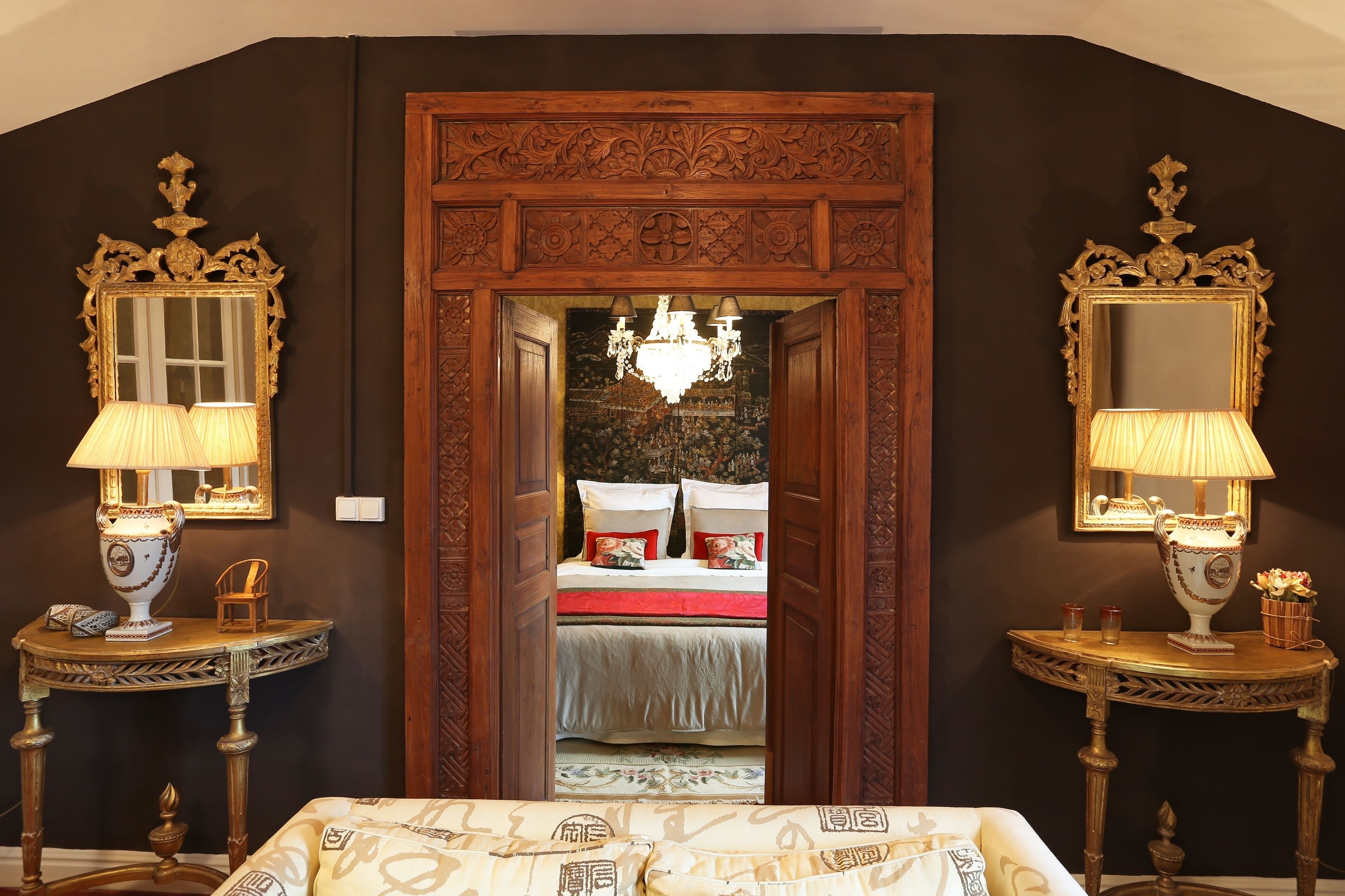 A room with a large ornate wooden door, two side tables with lamps, and a bed with a red and white bedspread.