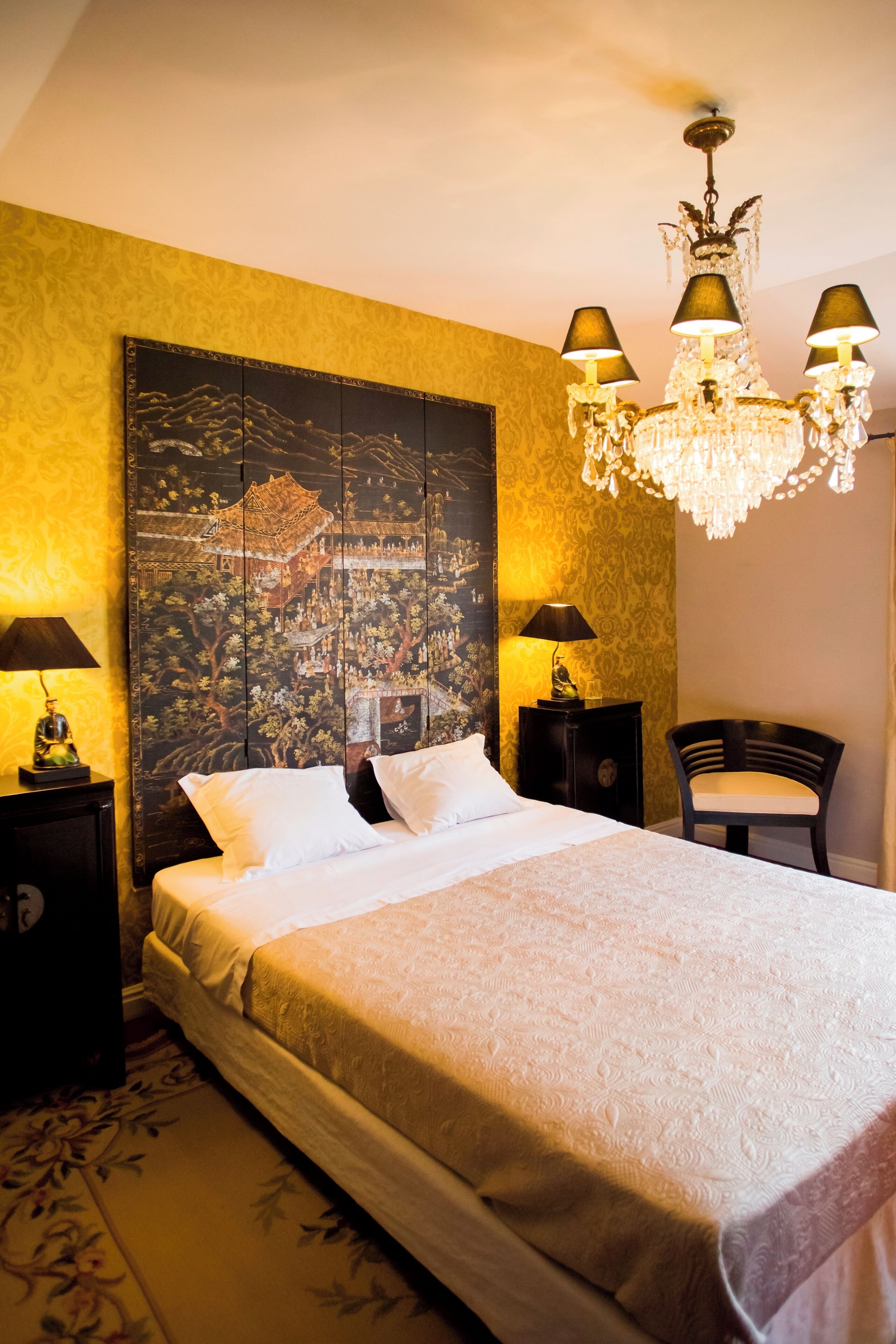 A neatly made bed with white linens, a large wall tapestry, a chandelier, and two bedside tables with lamps.