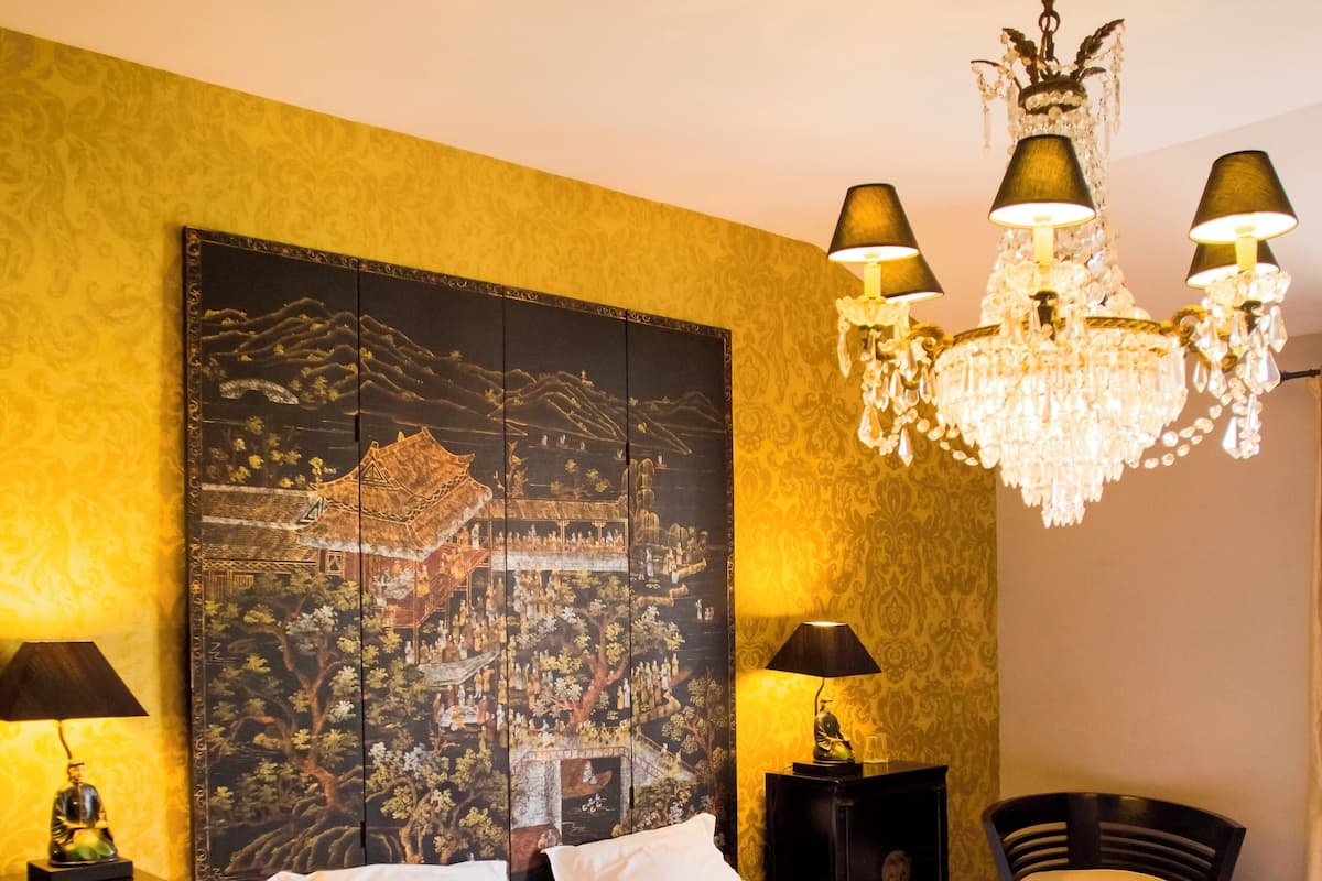A neatly made bed with white linens, a large wall tapestry, a chandelier, and two bedside tables with lamps.