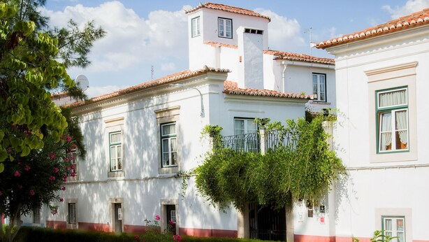 A white building with red trim and a tiled roof, surrounded by greenery and purple flowers. - Casa do Terreiro do Poço (Borba)