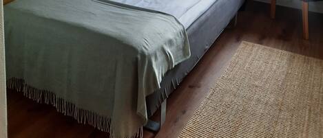 Un dormitorio con cama, escritorio, silla, lámpara, ventana con cortinas, planta y un cuadro en la pared.