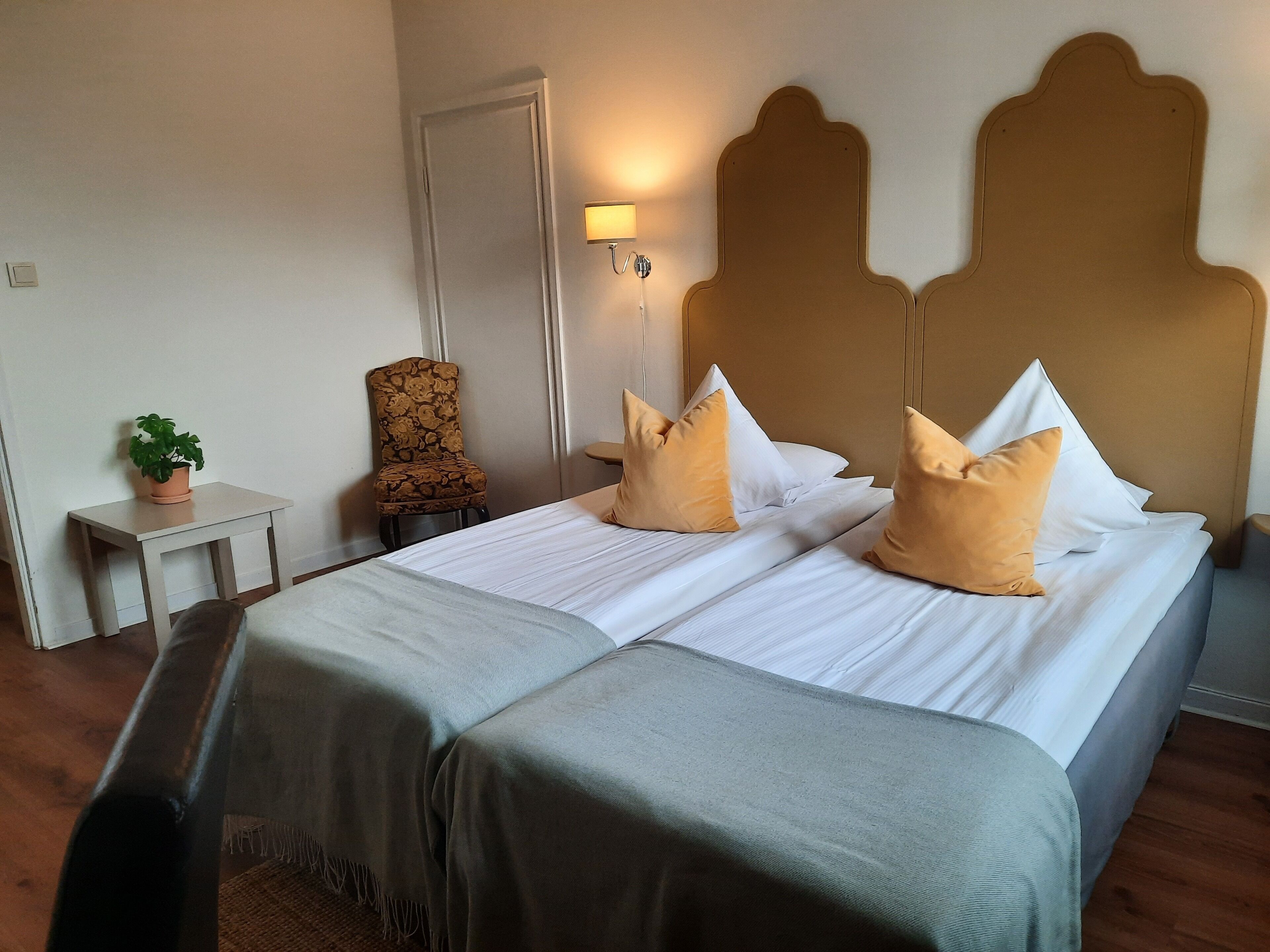 A hotel room with a bed, two pillows, a chair, a small table with a plant, and a wall-mounted lamp.