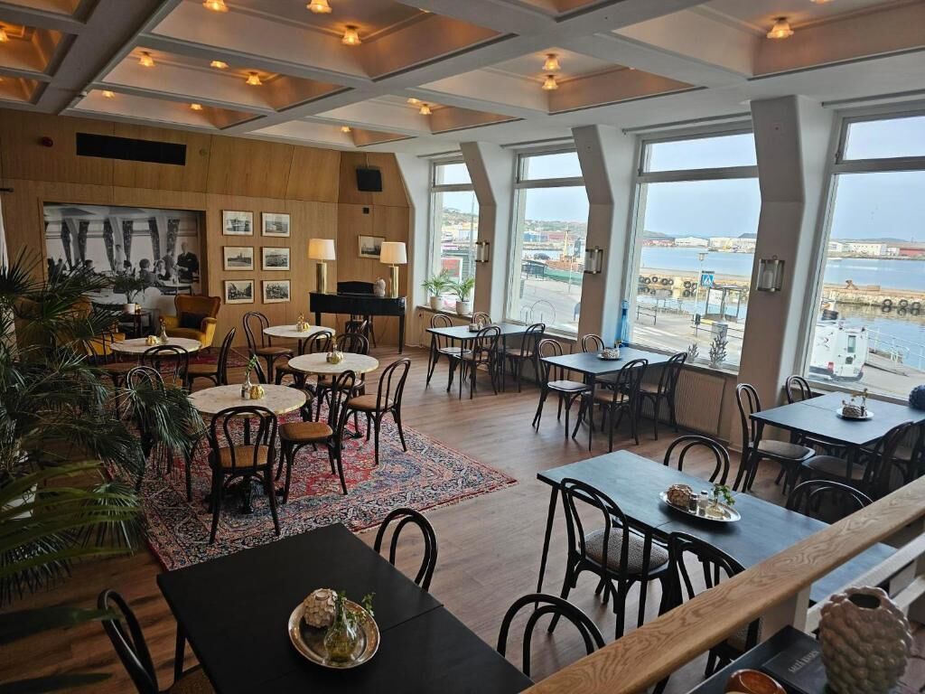 A restaurant with multiple tables and chairs, large windows with a view of the sea, and a wooden ceiling.