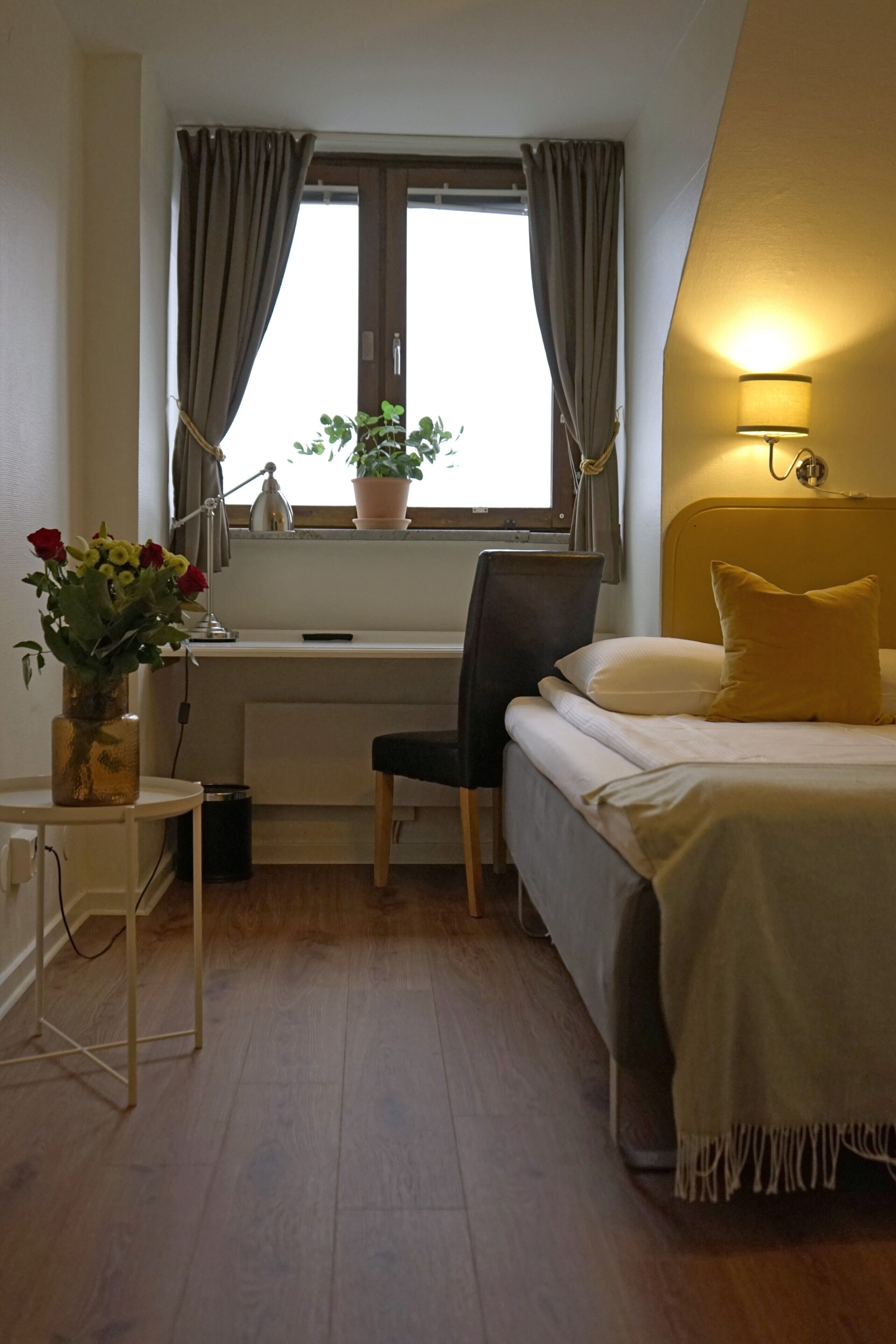 Een slaapkamer met een bed, een bureau, een stoel, een kamerplant en een lamp.