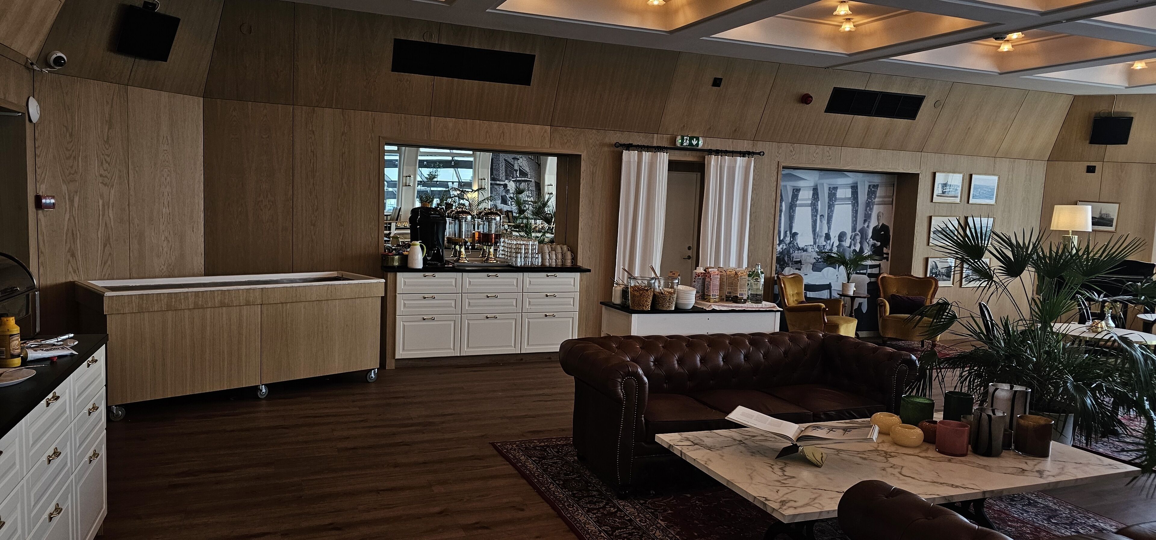 A modern hotel lobby with a wooden floor, a leather sofa, a marble coffee table, and a kitchen area with cabinets and appliances.