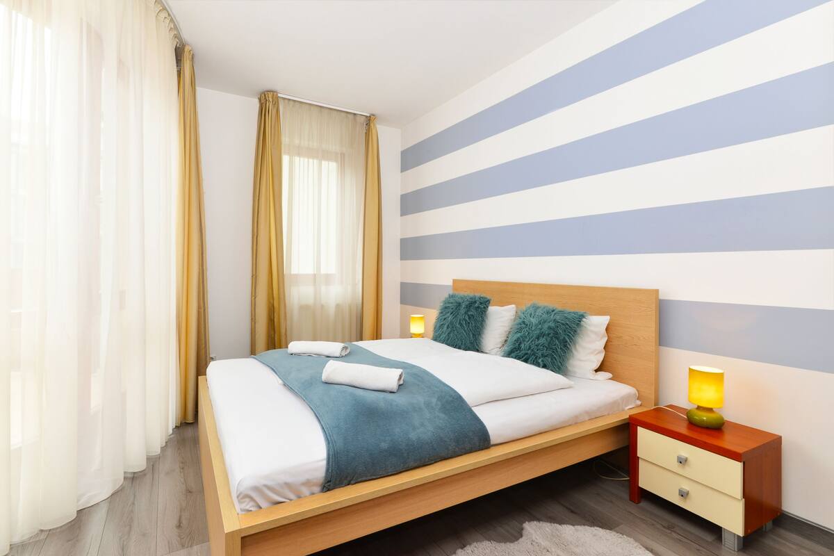 A modern bedroom with a wooden bed, a nightstand with a lamp, and a striped wall.