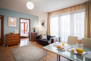 Una habitación de hotel moderna con zona de comedor, sofá, mesa de centro y vistas a la ciudad a través de grandes ventanales.