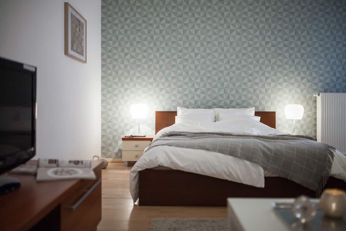 A neatly arranged bedroom with a bed, bedside lamps, a desk with a computer, and a framed picture on the wall.