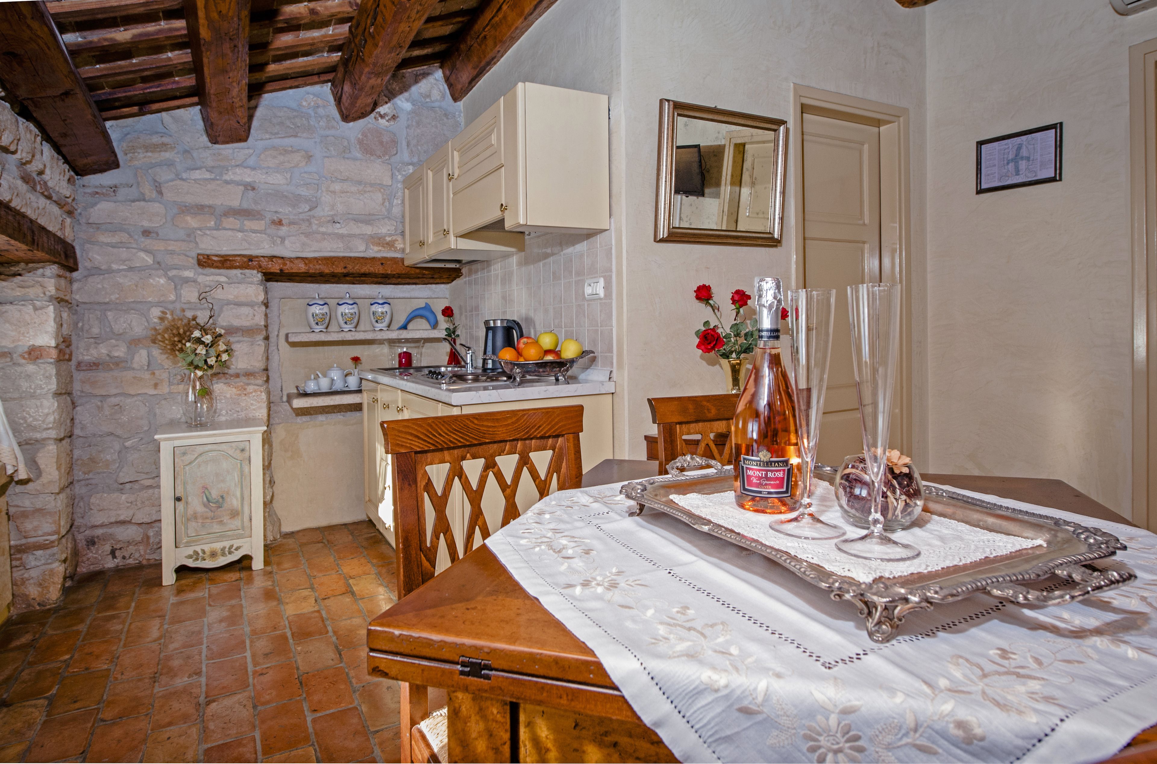 Une cuisine rustique aux murs de pierre, avec des poutres en bois et une table à manger agrémentée d’une bouteille, de verres et d’un panier de fruits.