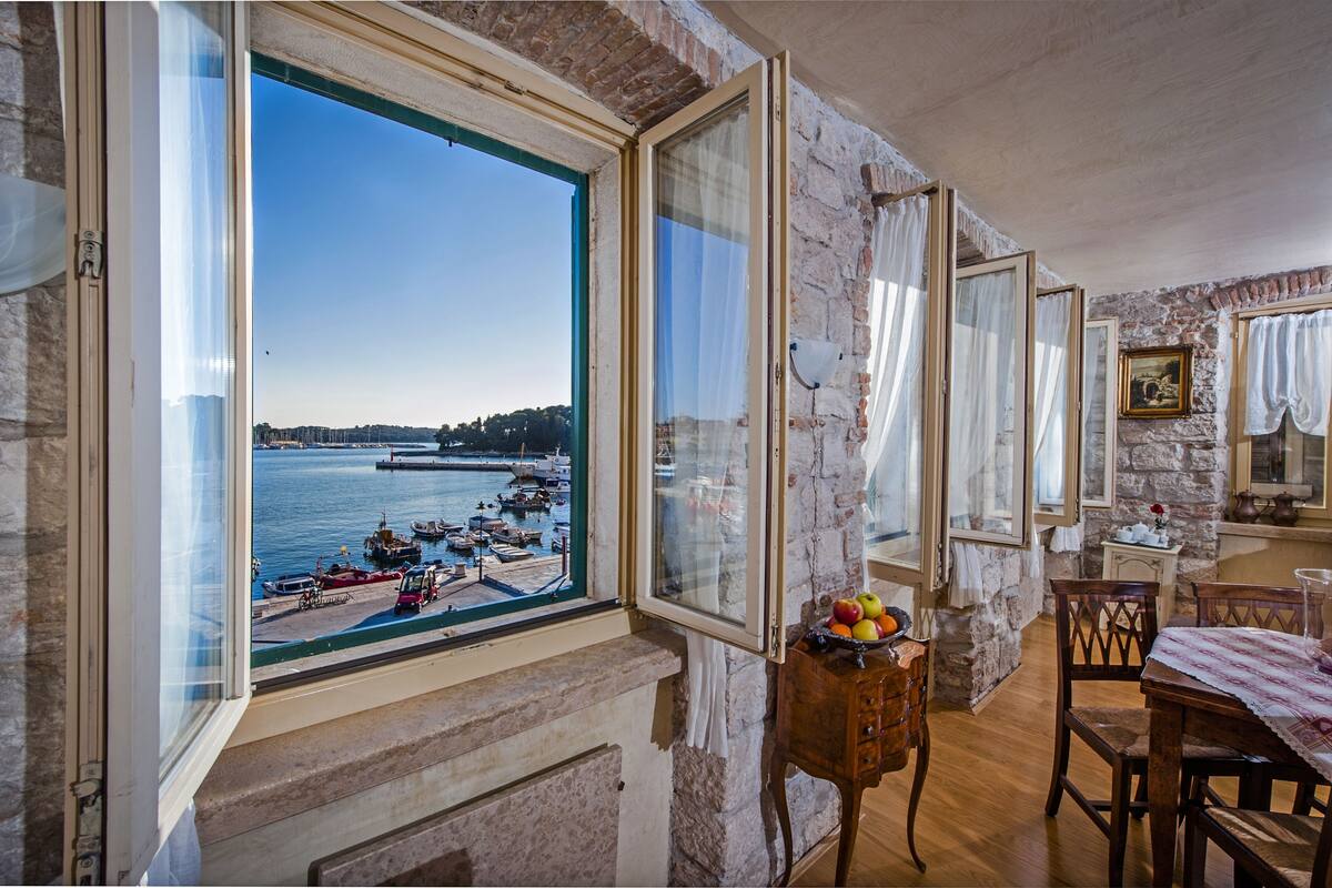Una habitación con un amplio ventanal con vistas al puerto, una mesa de madera con sillas y un pequeño mueble con fruta.