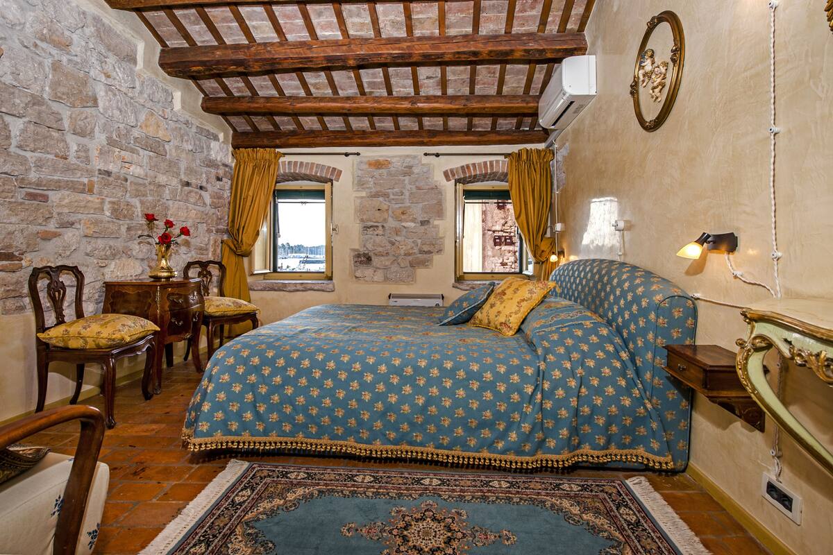 Un dormitorio con pared de piedra, vigas de madera en el techo, una cama con colcha estampada, una mesita de noche, una lámpara y vistas a través de una ventana.
