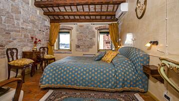 A bedroom with a stone wall, wooden ceiling beams, a bed with a patterned bedspread, a nightstand, a lamp, and a view through a window.
