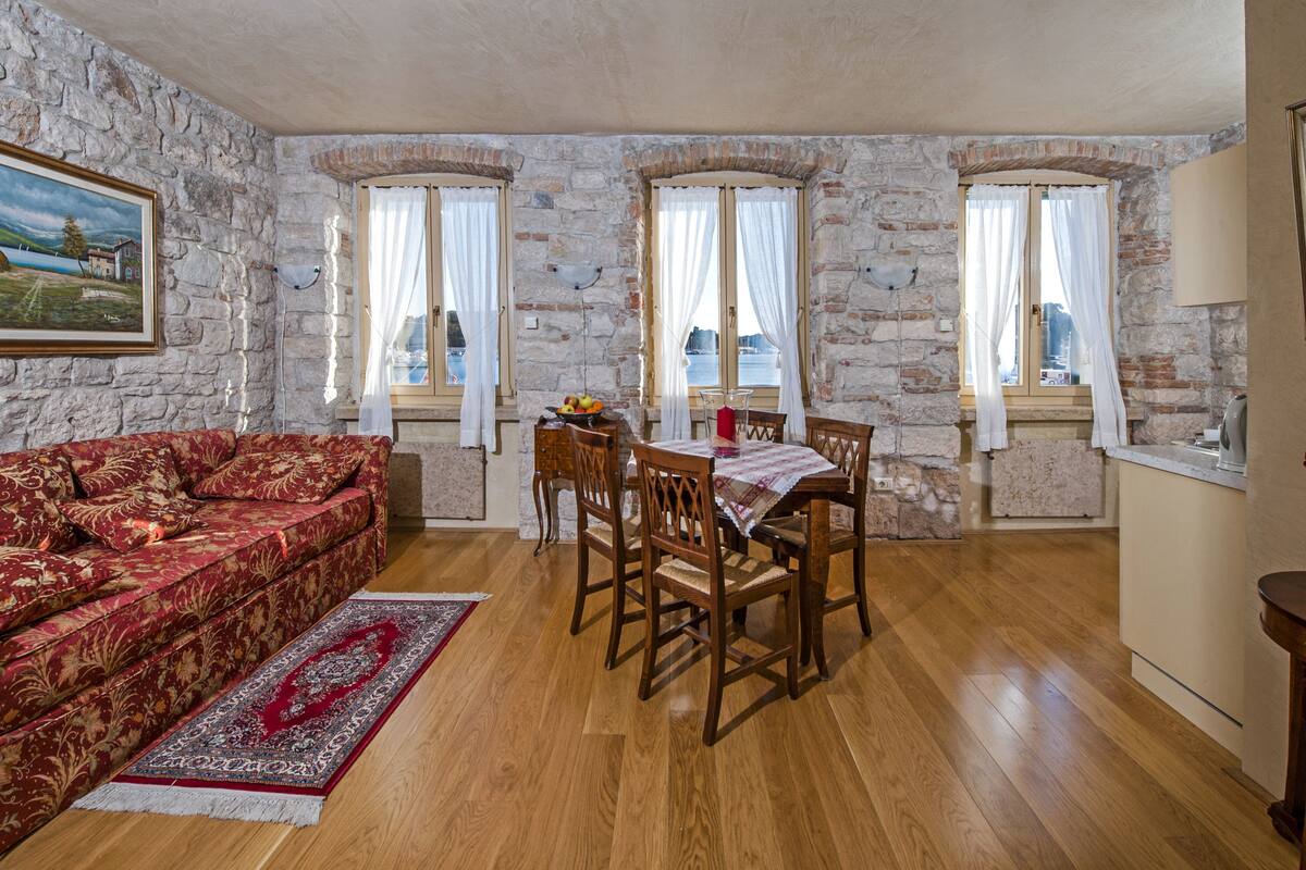 Una habitación con pared de piedra, un sofá rojo estampado, una mesa de comedor de madera con sillas y vistas al mar a través de ventanas.