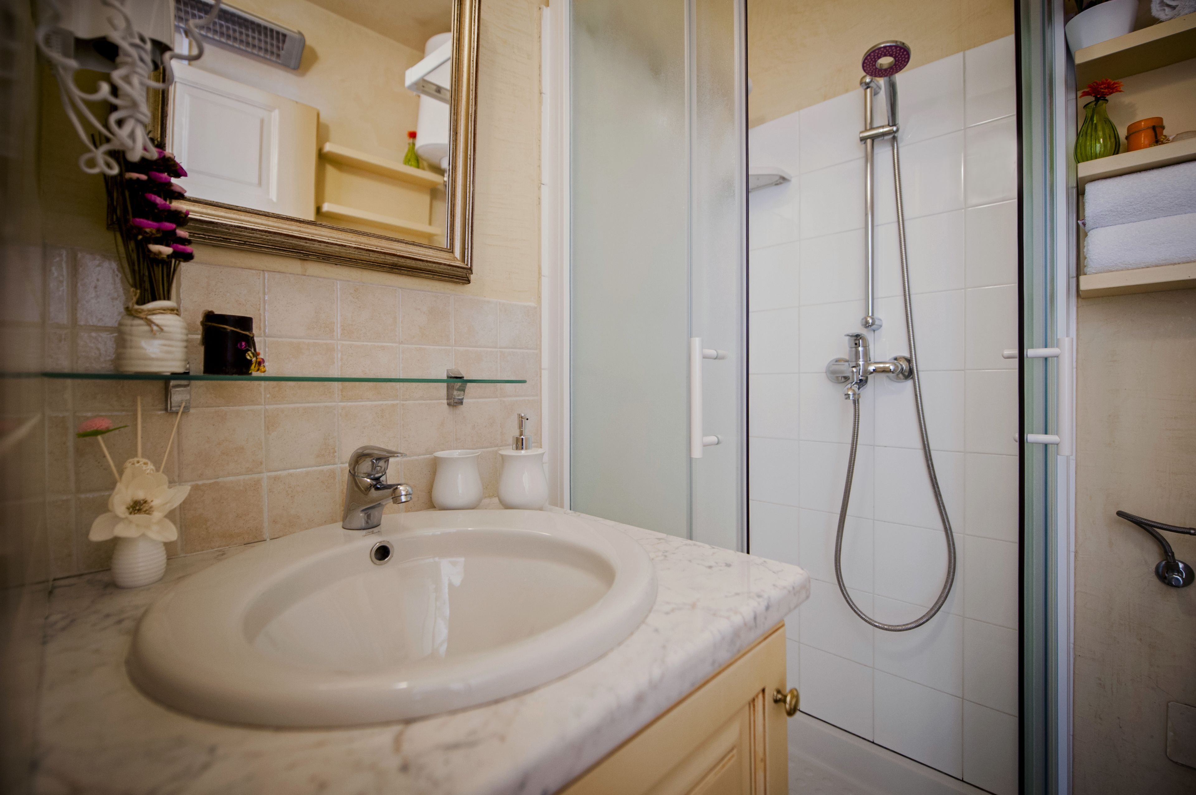 Een badkamer met een wastafel, spiegel, douche en glazen planken met toiletartikelen.