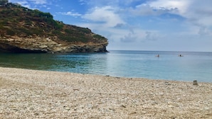 Une plage de galets bordée d’une eau calme, une falaise rocheuse et, au loin, des personnes pratiquant le kayak.