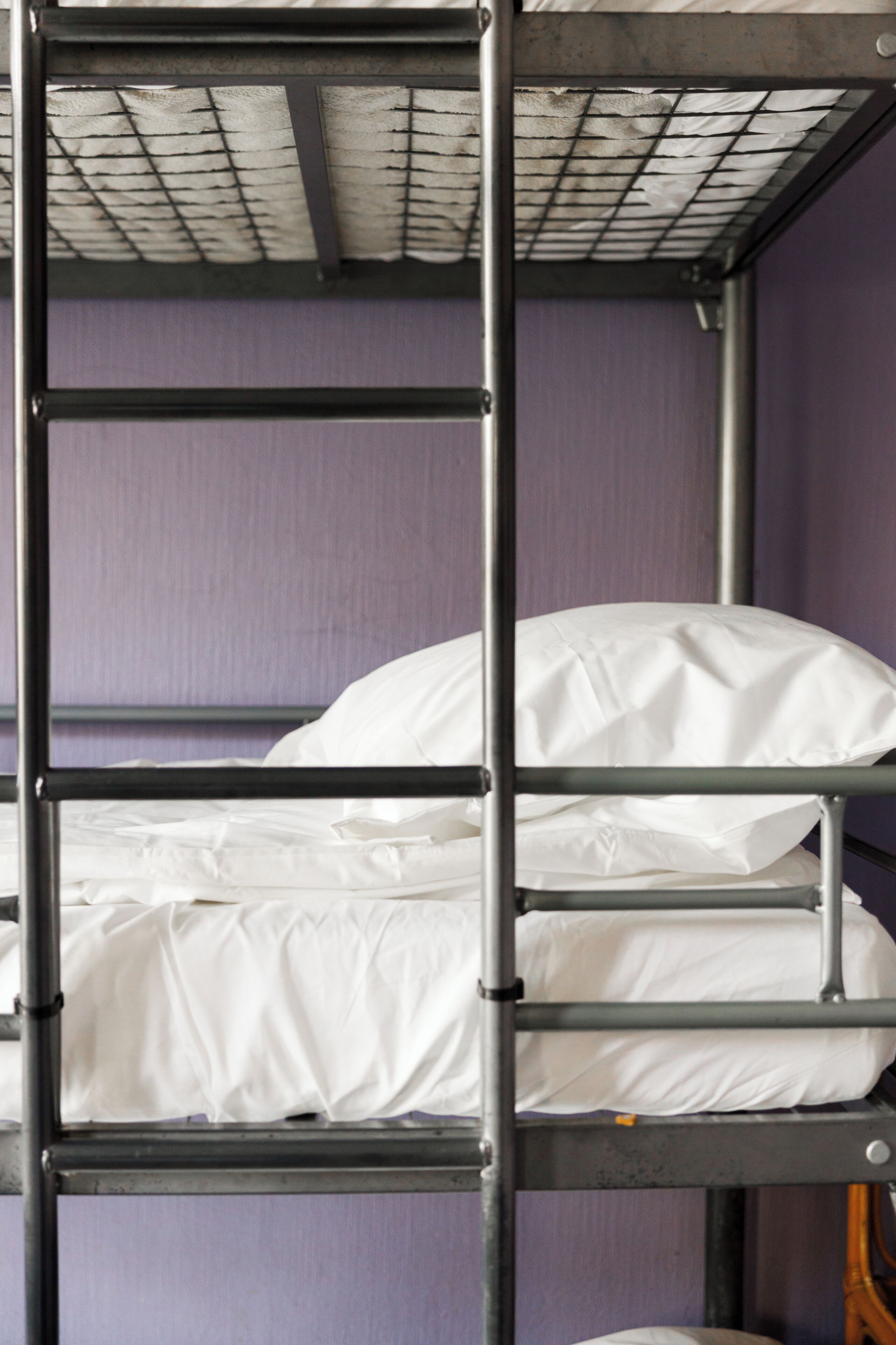 Una cama litera con ropa de cama blanca, barandales de metal y una pared morada.